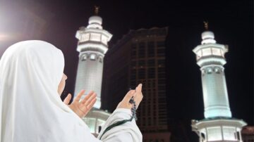 امرأة ترتدي عباءة بيضاء تدعو بإخلاص أمام مسجد الحرم في مكة المكرمة، مع أبراج المسجد مضاءة في الليل.
