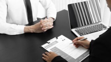 شخص يجلس أمام جهاز كمبيوتر محمول أثناء إجراء مقابلة عمل، بينما يقوم الآخر بكتابة معلومات على استمارة سيرة ذاتية. يظهر التركيز والاحترافية في بيئة مكتبية.