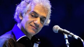 مغني مشهور يقف أمام ميكروفون، مبتسم، مع خلفية زرقاء مضاءة. يتمتع بشعر فضي ومظهر أنيق، يرتدي قميصًا أسود مع تفاصيل بيضاء.