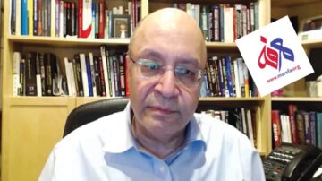 رجل يجلس أمام مكتبة تحتوي على العديد من الكتب، يظهر تعبيره الجاد. في الزاوية العلوية اليمنى، يوجد شعار لموقع إلكتروني.