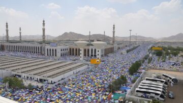 تجمع حشود كبيرة من الحجاج في مشعر عرفات خلال موسم الحج، مع رؤية واضحة للمسجد والمرافق المحيطة. تظهر الأعلام والقبعات الملونة تحت أشعة الشمس، بينما يحيط الجبال بالمنطقة.