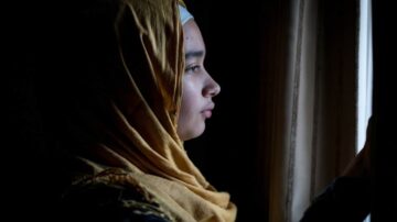 فتاة ترتدي حجابًا ذهبيًا تنظر من نافذة في غرفة مظلمة، الإضاءة تسلط الضوء على جانب وجهها.