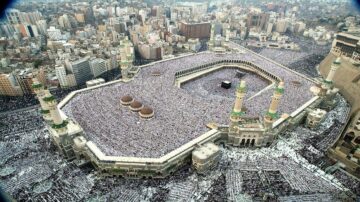 صورة جوية للحرم المكي في مكة المكرمة، تظهر الكعبة المشرفة محاطة بحشود كبيرة من الحجاج. المدينة الحديثة تحيط بالمسجد، بينما يتوزع الناس داخل الساحة. تعكس الصورة روحانية المكان وأهمية الحج في الإسلام.