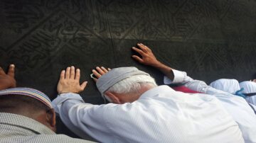 أشخاص يؤدون العمرة أو الحج، يلمسون الكعبة المشرفة، يظهرون من الخلف وهم يضعون أيديهم على الجدار الأسود للكعبة، في مشهد يعكس الروحانية والتقرب إلى الله.