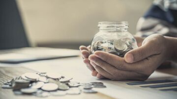 يد تحمل جرة زجاجية مليئة بالنقود المعدنية، مع انتشار بعض العملات على سطح المكتب. يظهر خلفية ضبابية تتضمن مستندات مالية. تعكس الصورة مفهوم الادخار وإدارة الأموال.