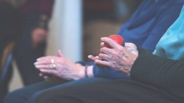 أيدٍ مسنّة تمسك بكرة حمراء، بينما تُظهر يد أخرى حركة التصفيق، في بيئة تجمع اجتماعي.