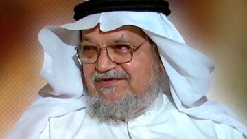 رجل مسن يرتدي الزي التقليدي العربي مع غترة وعقال، يظهر بابتسامة خفيفة، مع خلفية ملونة ناعمة.