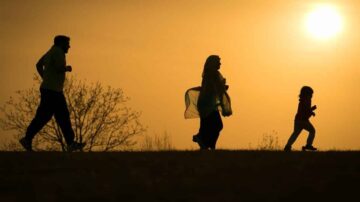 صورة ظلية لعائلة تتنزه في غروب الشمس. يظهر شخصان يمارسان الجري، بينما تسير امرأة مع طفل. الخلفية تظهر شجرة عارية وأشعة الشمس الذهبية. تعكس الأجواء النشاط والترابط العائلي.