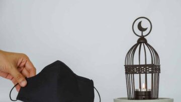 قناع وجه أسود يُمسك بيد بالقرب من فانوس معدني يحمل شمعة مضاءة، مع خلفية بيضاء بسيطة، يُظهر مزيجًا من عناصر العناية بالصحة والديكور المنزلي.