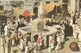 ساحة مزدحمة في سوق تقليدي، حيث يتجمع الناس حول نافورة حجرية. تظهر الخيام والسلع المعروضة، بينما يرتدي الزوار ملابس تقليدية. تعكس الصورة الحياة اليومية في السوق وتنوع الثقافات.