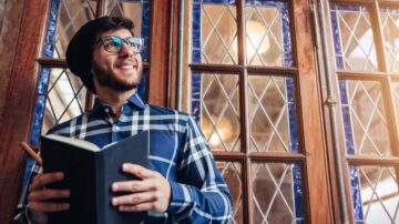 رجل يرتدي نظارات وقميص مزخرف يقف أمام نافذة زجاجية ملونة، مبتسمًا وهو يحمل دفتر ملاحظات وقلم. تعكس الإضاءة الطبيعية في الخلفية أجواءً دافئة ومريحة.
