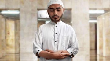 شاب يرتدي قميصًا أبيض وقبعة تقليدية، يقف في مكان داخلي مع جدران حجرية، يبدو في حالة تأمل أو صلاة.