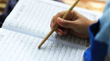 يد تحمل قلمًا خشبيًا وتكتب في صفحات كتاب يحتوي على نصوص عربية، مع التركيز على تفاصيل الكتابة.