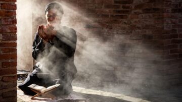 رجل يصلي في مكان مضاء بأشعة الشمس، جالسًا على الأرض مع كتاب مفتوح أمامه، محاطًا بالضباب. يعبر عن لحظة تأمل روحي وعبادة في بيئة هادئة.