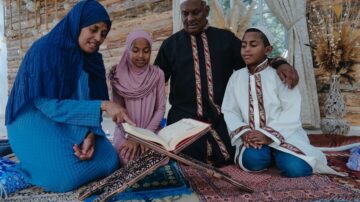 عائلة مسلمة تتجمع حول مصحف في مكان مريح. الأم ترتدي لباس تقليدي وتقرأ، بينما الأطفال يستمعون بتركيز. الأجواء تعكس الانتماء الثقافي والروحانية.
