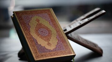 كتاب قرآن كريم مفتوح على حامل خشبي مع خلفية ضبابية. تصميم غلاف الكتاب مزخرف بألوان دافئة ونقوش دقيقة.