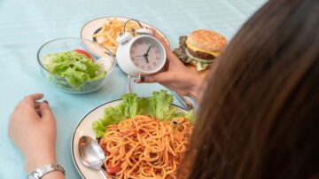 امرأة تحمل ساعة توقيت في يدها، بينما تحتوي الطاولة على طبق مكرونة مع صلصة، سلطة خضراء، ووجبة برغر مع بطاطس مقلية. تُظهر الصورة أهمية التوقيت في تناول الوجبات الصحية.