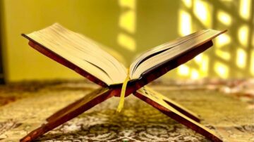 كتاب مفتوح موضوع على حامل، مع أشعة الشمس تتسرب من النوافذ خلفه، مما يخلق ظلالًا جميلة على السجادة.