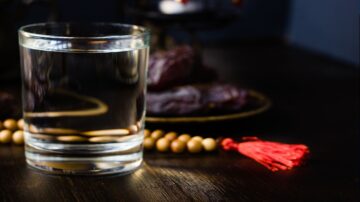 كوب زجاجي يحتوي على ماء صافٍ، موضوعة على طاولة خشبية داكنة، مع خلفية غير واضحة تحتوي على تمر ومسبحة خشبية.