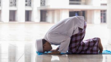 شاب مسلم يؤدي الصلاة في مسجد، يركع على سجادة صلاة زرقاء، مع خلفية معمارية حديثة.