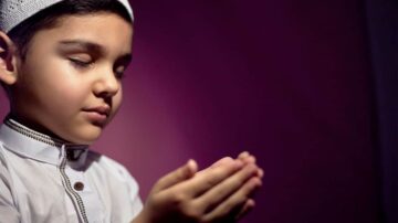 طفل صغير يرتدي زيًا تقليديًا، يتمتم بدعاء بيديه المرفوعتين، مع إغلاق عينيه في لحظة من الخشوع والتأمل. الخلفية مائلة للون الأرجواني، مما يضيف جوًا من السكينة والروحانية.