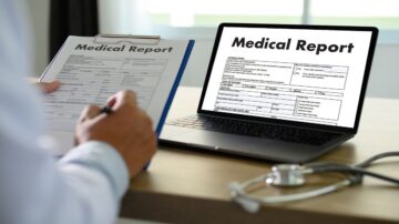 تقرير طبي معروض على شاشة كمبيوتر محمول، بينما شخص يحمل ملف يحتوي على تقرير طبي مكتوب. يظهر جهاز طبي على الطاولة.