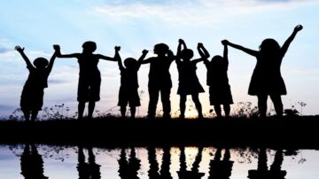 أطفال يقفون في صف على حافة، يرفعون أيديهم نحو السماء، silhouetted ضد غروب الشمس. تعكس المياه الهادئة أمامهم صورهم، مما يضيف لمسة جمالية للمشهد.