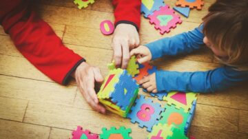 طفل صغير يجلس على الأرض ويلعب بألعاب تركيب ملونة مع شخص بالغ، حيث يضعان قطعاً من الأرقام والحروف على سجادة خشبية.