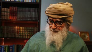 رجل مسن ذو لحية وشعر أبيض، يرتدي عمامة وقميصًا أخضر، يجلس في مكتبة تحتوي على كتب مرتبة. يظهر التركيز والتفكير العميق في تعبير وجهه.