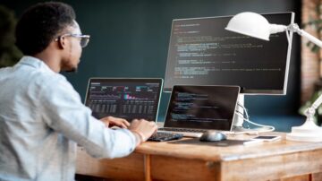 رجل يعمل على عدة شاشات كمبيوتر، مع وجود رموز برمجية ورسوم بيانية تظهر على الشاشات. يوجد لابتوب آخر على الطاولة مع لوحة مفاتيح وفأرة وهاتف محمول. الأجواء تعكس بيئة عمل تقنية حديثة.