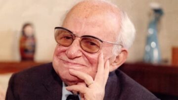 رجل مسن يرتدي نظارات وقميص رسمي، يجلس مبتسمًا ويضع يده على ذقنه. الخلفية تحتوي على عناصر منزلية بسيطة، مما يضفي جوًا وديًا.