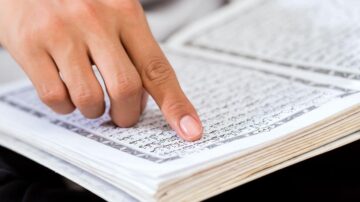 يد تشير إلى نص مكتوب بخط عربي في كتاب مفتوح، مع التركيز على تفاصيل الصفحة.