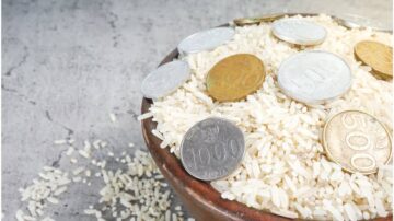 وعاء من الأرز الأبيض مع عملات معدنية متنوعة موضوعة فوقه وعلى الأرضية المحيطة. الأرز يظهر بشكل وفير، بينما العملات تشمل فئات مختلفة تعكس قيمة مالية.