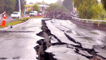 صورة تظهر طريقًا متصدعًا نتيجة زلزال، مع انهيار جزئي للأرض. تظهر على الجانبين علامات تحذيرية وأقماع مرورية، مما يشير إلى منطقة غير آمنة. المنظر يوضح الأضرار التي لحقت بالبنية التحتية.