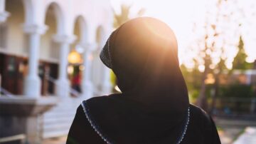 امرأة ترتدي حجابًا أسود تقف في ضوء الشمس، مع خلفية تضم مبنى ذو قوس، مما يخلق أجواء هادئة وجميلة.