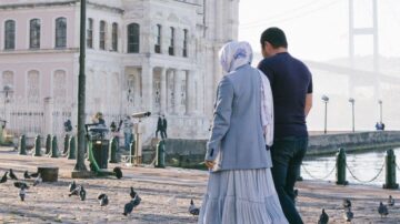 رجل وامرأة يسيران معًا على ضفاف المياه، حيث تحلق الحمام حولهما. المباني التاريخية تظهر في الخلفية مع جسر يلوح في الأفق. الجو هادئ ومشمس، مما يضفي طابعًا رومانسيًا على المشهد.