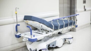 سرير طبي متنقل بلون أزرق، موضوع بجانب جدار في ممر مستشفى. السرير مزود بعجلات ويدعم نظام رفع، مع وجود أنبوب متصل.