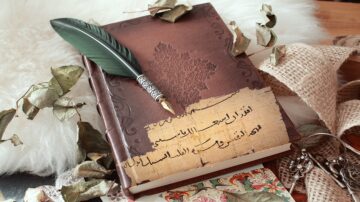 كتاب قديم مزخرف مع قلم ريش أخضر، يظهر فوقه أوراق جافة. الكتاب يحتوي على نص عربي قديم. يحيط بالكتاب قماش محاكة وأوراق ملونة، مما يضيف لمسة فنية للمشهد.