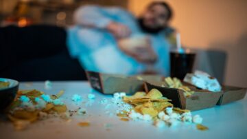 رجل مسترخٍ على الأريكة يتناول الوجبات الخفيفة، مع أكواب من رقائق البطاطس والفشار متناثرة على الطاولة. الخلفية ضبابية، ويظهر مشروب غازي بجانب الطعام.