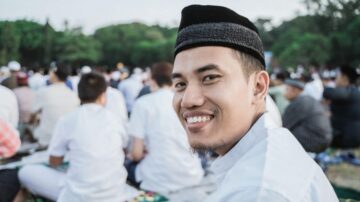 رجل مبتسم يرتدي زيًا تقليديًا في تجمع لمناسبة دينية في الهواء الطلق، مع خلفية من الحضور في الحديقة.