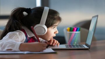فتاة صغيرة ترتدي سماعات رأس بيضاء، تركز على شاشة الكمبيوتر المحمول أثناء الدراسة. أمامها دفتر وقلم، مع حاوية ملونة تحتوي على أقلام. تعكس الصورة بيئة تعليمية منزلية.