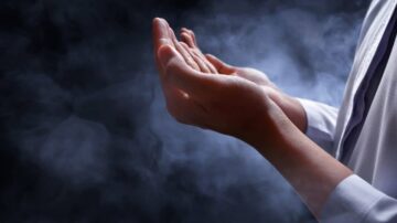 يدان مرفوعتان في الضوء الخافت مع دخان يتصاعد حولهما، تعبير عن التأمل أو الرغبة في الشفاء.