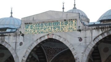 خطوط عربية مزخرفة على واجهة مسجد، تتضمن آيات قرآنية، مع قبب مميزة. تعكس العمارة الإسلامية التقليدية.