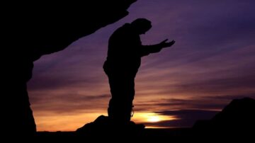 شخص يقف في ظل صخرة، يرفع يديه نحو السماء عند غروب الشمس. الألوان تتدرج من البرتقالي إلى البنفسجي، مما يخلق جوًا هادئًا وروحيًا.