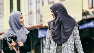 امرأتان ترتديان الحجاب، واحدة تحمل كاميرا، يتبادلان الابتسامات في منطقة حضرية. الخلفية تحتوي على مبانٍ ملونة وأجواء مبهجة.