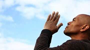 رجل يرفع يديه نحو السماء في وضعية دعاء، مع خلفية سماء زرقاء مغطاة بكتل من السحاب. تعبر الصورة عن لحظة من التأمل والروحانية.