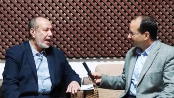 رجلان يجلسان في غرفة ذات تصميم داخلي بسيط. الرجل على اليسار يرتدي بدلة داكنة ويبدو أنه يتحدث، بينما الرجل على اليمين يرتدي سترة رمادية ويحمل ميكروفون. الخلفية عبارة عن جدار مزخرف بشبكة خشبية.