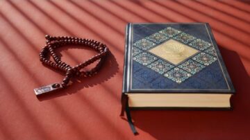 مصحف القرآن الكريم بجوار سبحة خشبية على سطح برتقالي. الضوء يتسلل عبر خطوط الظل، مما يبرز تفاصيل الغلاف المزخرف للقرآن.