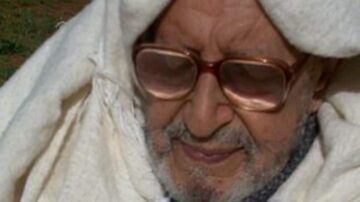 رجل مسن يرتدي غطاء رأس أبيض، يجلس في الهواء الطلق، يظهر عليه الحكمة والهدوء. تفاصيل وجهه تعكس تجارب الحياة.