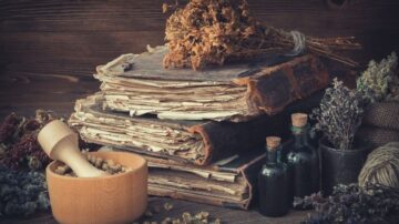 كتب قديمة متراكمة فوق بعضها، مع مجموعة من الأعشاب المجففة في الخلفية. يوجد وعاء خشبي يحتوي على مكونات طبيعية، وزجاجات صغيرة ملونة بجانب الكتب. تعكس الصورة جوًا من التراث والمعرفة في مجال الأعشاب والعلاج الطبيعي.